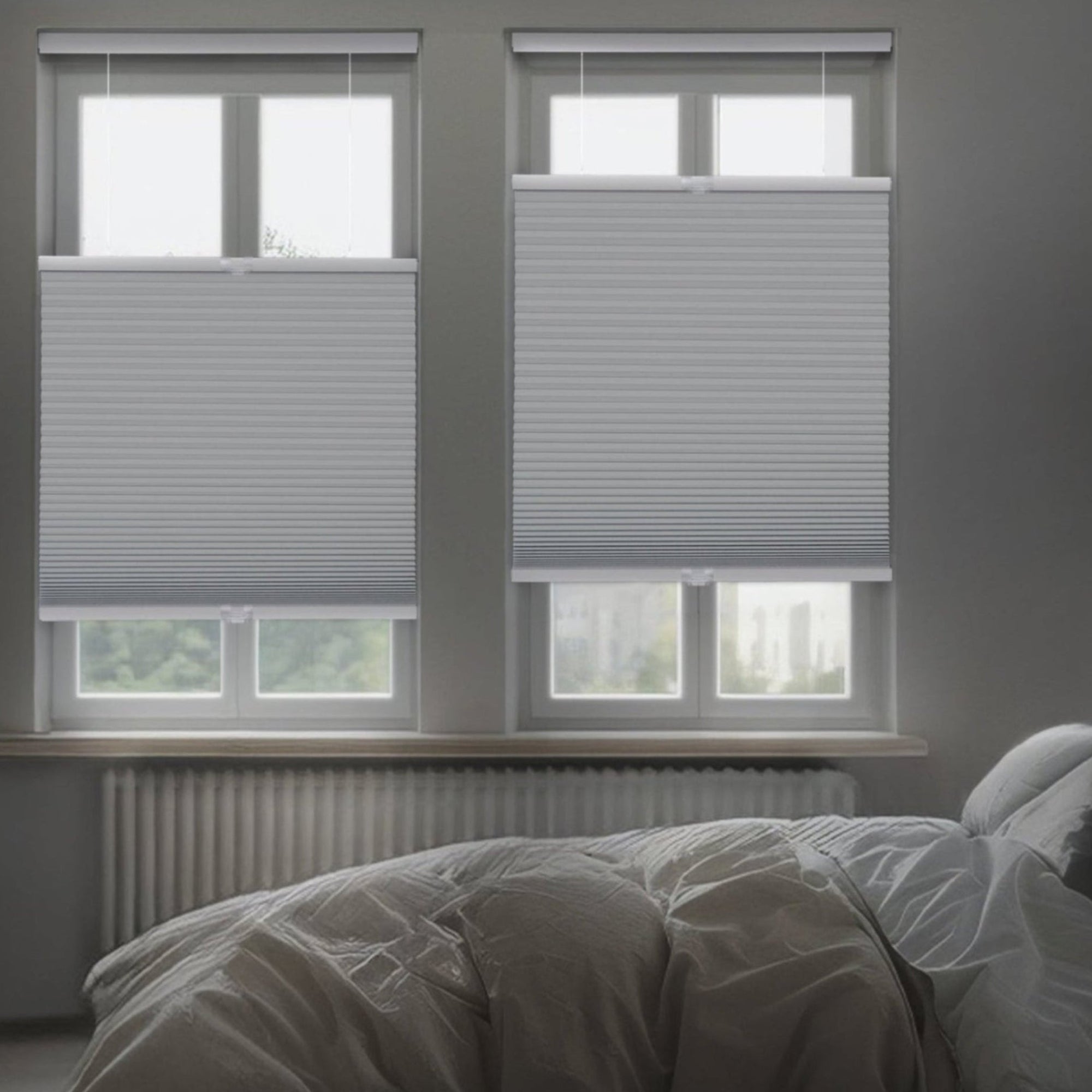 A dimly lit, modern bedroom featuring two gray top-down bottom-up honeycomb shades installed on side-by-side windows. The shades are partially lowered from the top to allow slivers of light while maintaining privacy over a bed with white and gray linens.