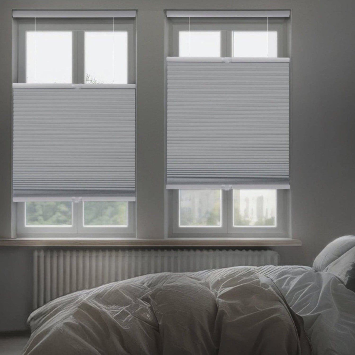 A dimly lit, modern bedroom featuring two gray top-down bottom-up honeycomb shades installed on side-by-side windows. The shades are partially lowered from the top to allow slivers of light while maintaining privacy over a bed with white and gray linens.