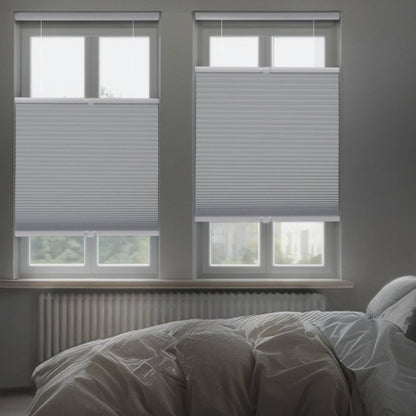 A dimly lit, modern bedroom featuring two gray top-down bottom-up honeycomb shades installed on side-by-side windows. The shades are partially lowered from the top to allow slivers of light while maintaining privacy over a bed with white and gray linens.