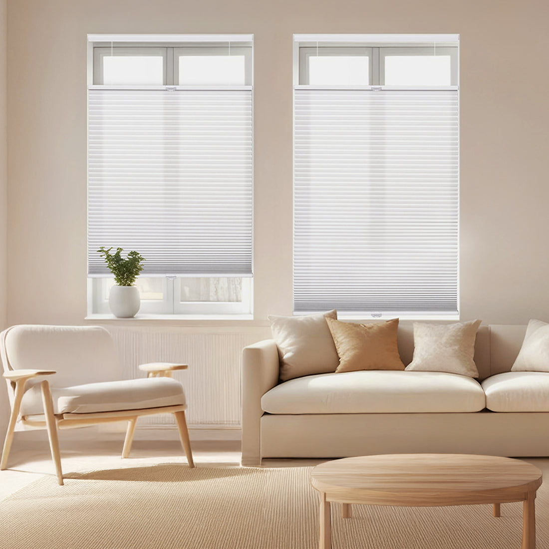 A bright, modern living room featuring two white cordless top-down bottom-up honeycomb shades installed on side-by-side windows. The minimalist space includes a cream-colored sofa with earth-tone pillows, a light wood armchair, and a small potted plant on the windowsill, all bathed in soft, natural light.